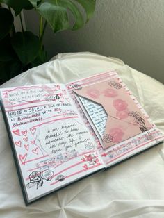 an open book with pink and white designs on it sitting on a bed next to a potted plant