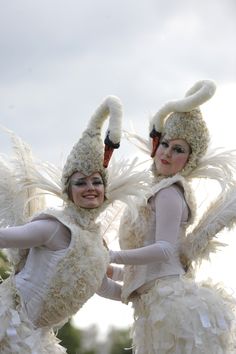 two women dressed in white costumes with feathers on their heads and arms, both holding hands behind their backs