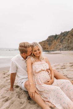 a pregnant woman sitting on the beach with her husband next to her and kissing her belly