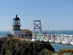 people walking across a bridge over the ocean with a lighthouse in the background on a sunny day