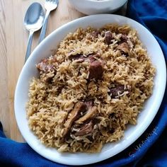 a white bowl filled with rice and meat on top of a wooden table next to a spoon