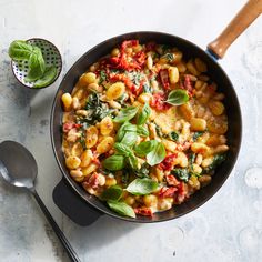 a skillet filled with pasta and spinach on top of a table next to a spoon