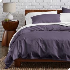 a bed with purple sheets and pillows in front of a white brick wall next to a night stand