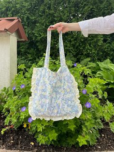 a hand holding a blue and white bag in front of green plants with purple flowers