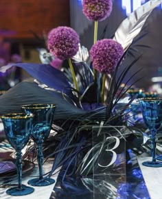 purple flowers are sitting on the table with blue glass goblets and silverware