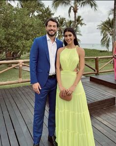 a man and woman in formal wear standing on a deck next to each other with palm trees behind them