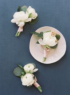 two white boutions and some green leaves on a blue tablecloth with a plate