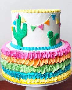 a multi - layered cake decorated with colorful icing and cactus decorations on a white pedestal