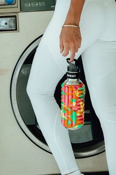 a woman in white leggings holding a water bottle filled with candy sticks next to a washing machine