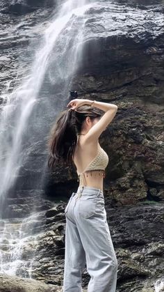 a woman standing in front of a waterfall with her arms behind her head, wearing high waist pants and crop top