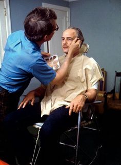 a man sitting in a chair getting his hair cut by an older gentleman who is standing next to him