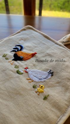 embroidered napkin with roosters on it sitting on top of a wooden table in front of a window