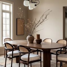 a dining room table with chairs and a vase on top of it in front of two windows