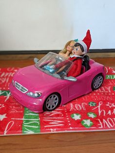 a pink toy car with an elf riding in the driver's seat on a christmas rug