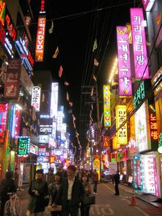 people walking down the street at night with neon signs on buildings and lights all around