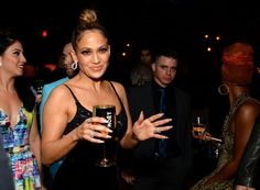 a woman in a black dress holding a wine glass with other people standing around her