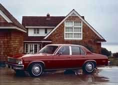 an old red car parked in front of a house on a wet driveway next to the ocean