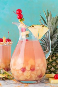 a pitcher filled with pineapple punch and garnished with cherries next to sliced pineapples