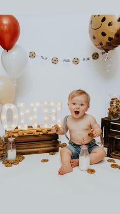 a baby boy sitting on the floor with milk and cookies in front of him, surrounded by balloons