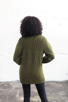 a woman standing in front of a white wall wearing a green sweater and black pants