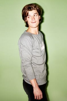 a young man standing in front of a green wall wearing a grey shirt and black pants