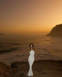 a woman standing on top of a rock next to the ocean at sunset with her arms behind her head