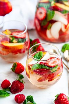 two glasses filled with fruit and ice on top of a white table next to spoons