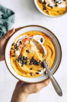 two hands holding a bowl of hummus with pumpkin sauce and sunflower seeds on top