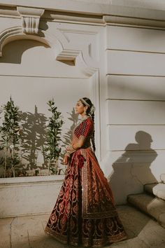 a woman standing in front of a white building wearing a red and gold lehenga