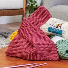 yarn and knitting needles are on the floor next to a red knitted bag that is sitting on a coffee table