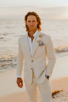 a man in a white suit standing on the beach with his hands in his pockets