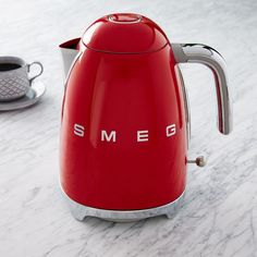 a red kettle sitting on top of a white counter next to a cup of coffee