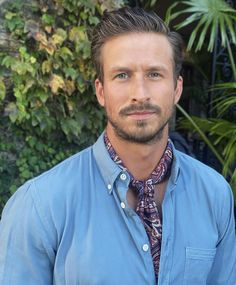 a man wearing a blue shirt and tie standing in front of some plants with his eyes closed