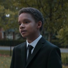 a young boy in a suit and tie looking at the camera with trees in the background