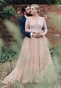 a man and woman standing next to each other in front of a brick wall wearing wedding gowns