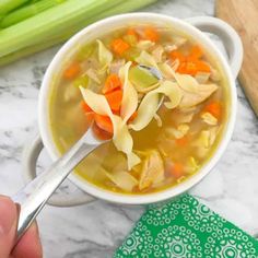 a person holding a spoon over a bowl of chicken noodle soup with celery and carrots