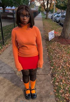 a woman in an orange sweater and red skirt stands on the sidewalk with her legs crossed