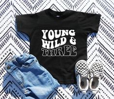 the young wild and three shirt is laying on top of some denim shorts, with shoes next to it