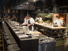 chefs preparing food in a restaurant kitchen with stainless steel counter tops and black chairs around them