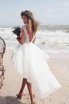 a woman in a white dress is walking on the sand near the ocean and holding flowers