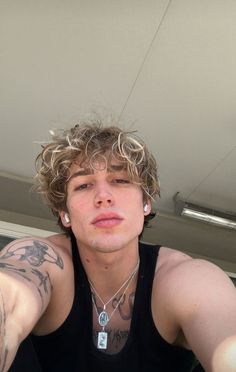 a young man with curly hair wearing a black tank top and silver necklace sitting in front of a window