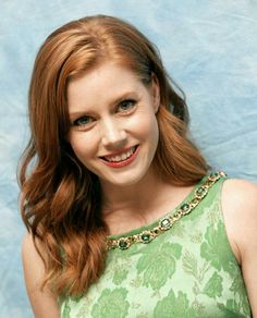 a woman with long red hair wearing a green dress and smiling at the camera while standing in front of a blue backdrop