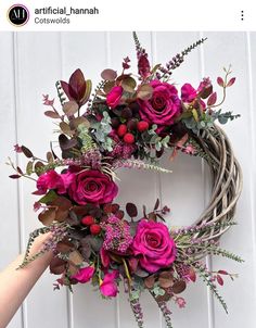 a wreath with pink flowers and greenery hanging on a white wooden door, being held by a person's hand