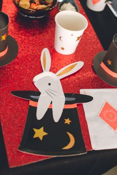 a table topped with paper hats and cups filled with food on top of red cloth