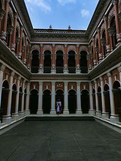 two people are standing in the middle of an empty courtyard with columns and arches on either side