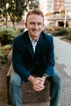 a man in a suit sitting on a bench smiling at the camera with his hands clasped