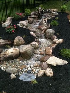 a garden with rocks and water flowing from it to the ground in front of a fence