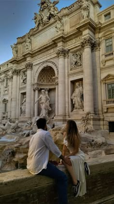 a man and woman sitting on the edge of a wall next to a fountain in front of a building