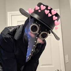 a man wearing a plague mask with pink hearts on his hat and goggles in front of a door