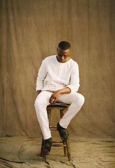 a man sitting on top of a wooden chair in front of a brown wall wearing white pants